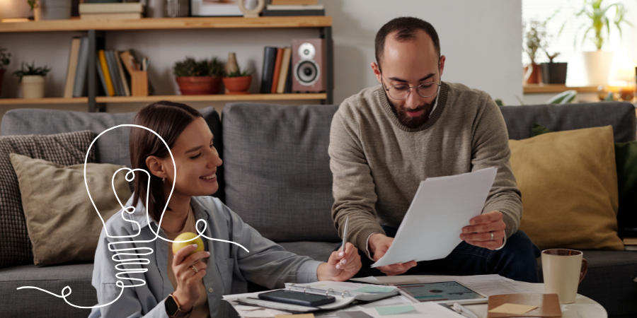 A couple sitting on a sofa reviewing household bills, smiling while going through paperwork together.